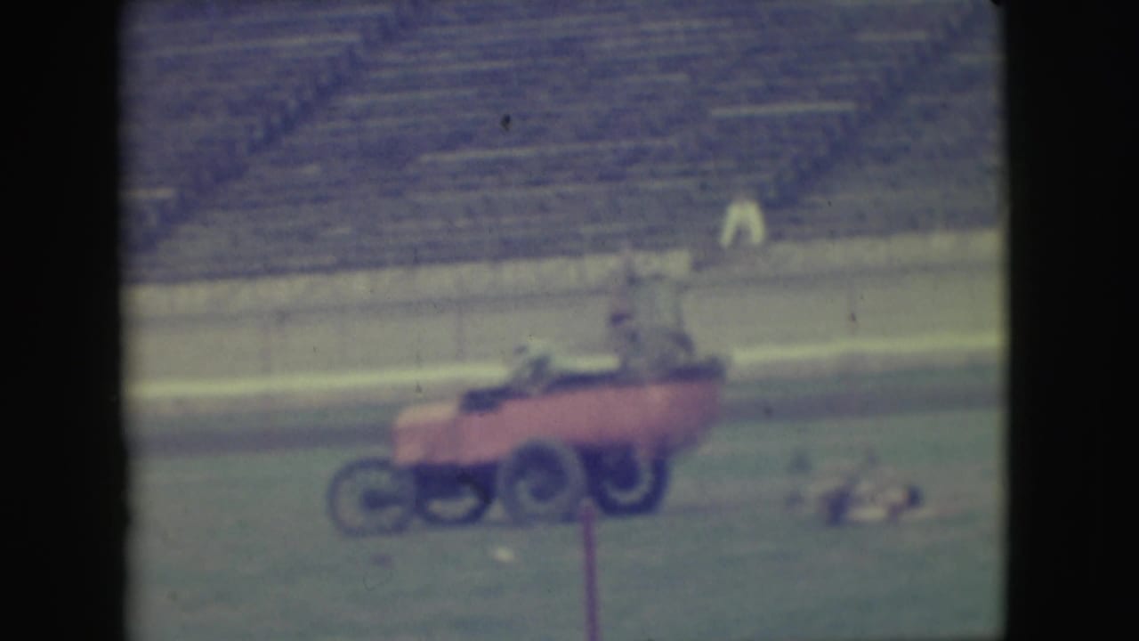 In 1949 Denver home-movie footage, three men appear to ride a red cart across a grassy field with empty bleachers behind them.