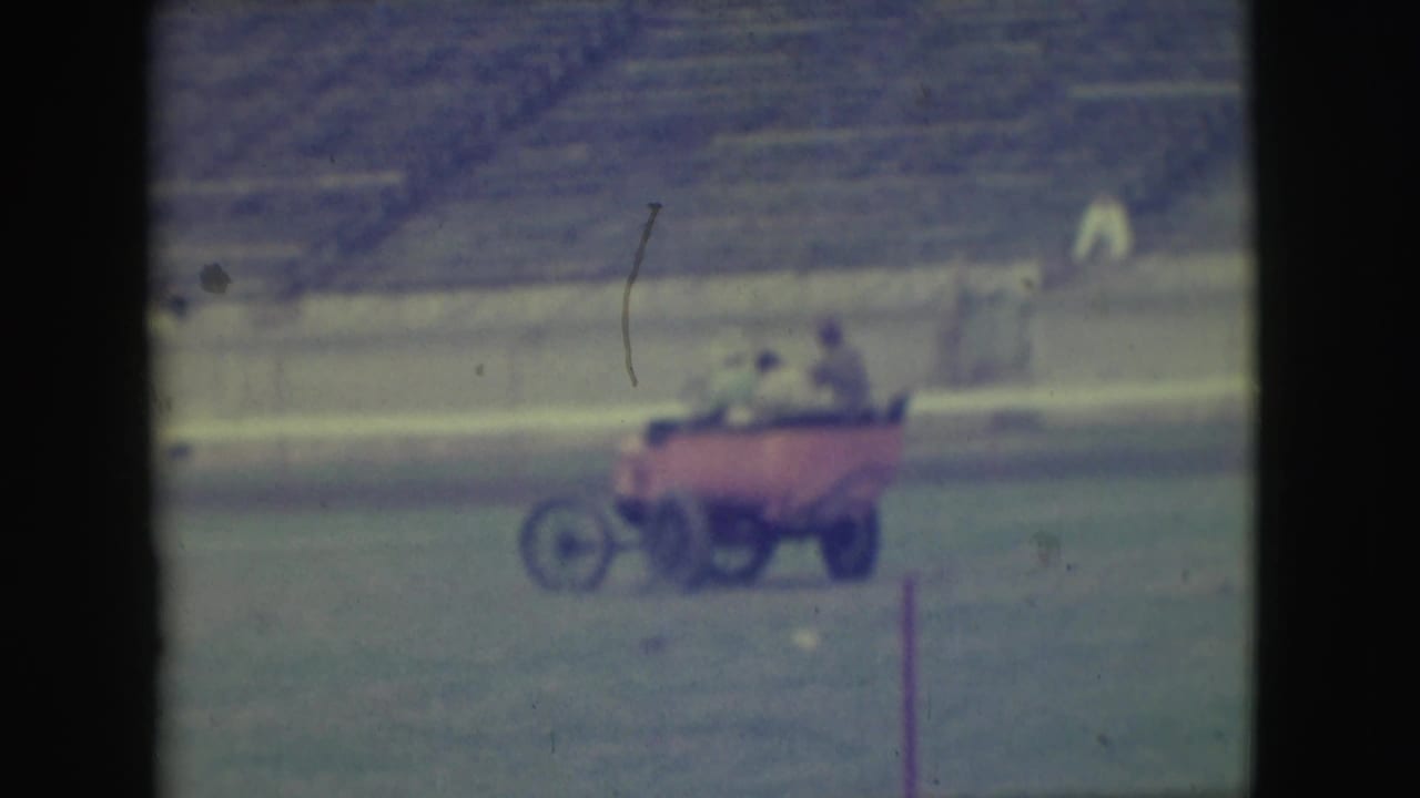 The red cart continues across the Denver stadium field, carrying several adult men with empty bleachers in the background.