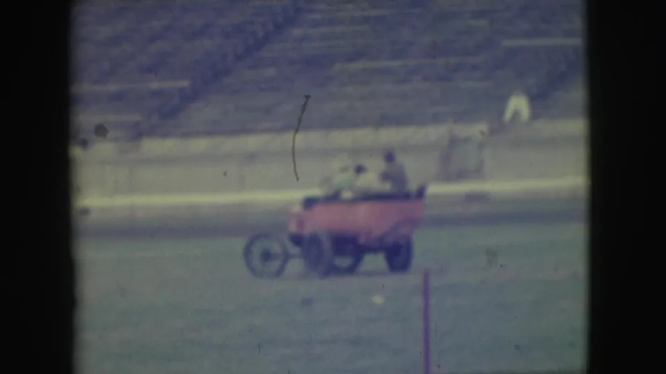The red cart continues across the Denver stadium field, carrying several adult men with empty bleachers in the background.