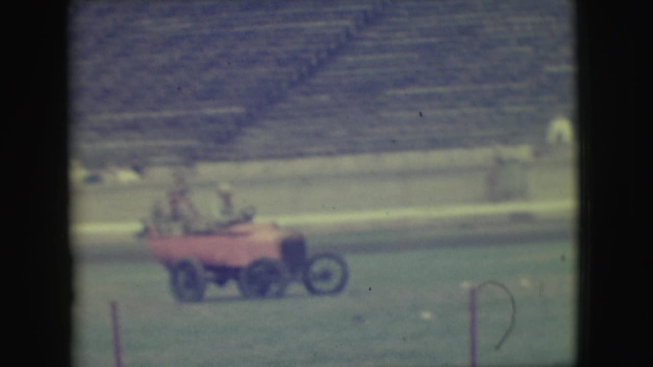 Red multi-wheel vehicle moving across a grassy field with riders and bleachers behind it.