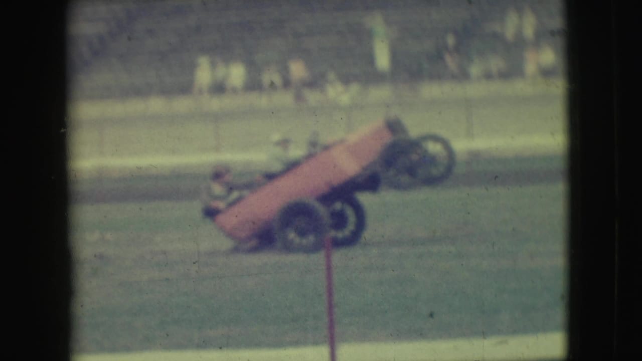 1949 Denver home-movie frame of an orange open-wheeled stunt car tipping onto two wheels before distant spectators.