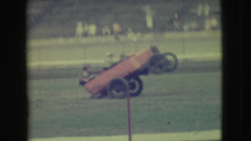 1949 Denver home-movie frame of an orange open-wheeled stunt car tipping onto two wheels before distant spectators.