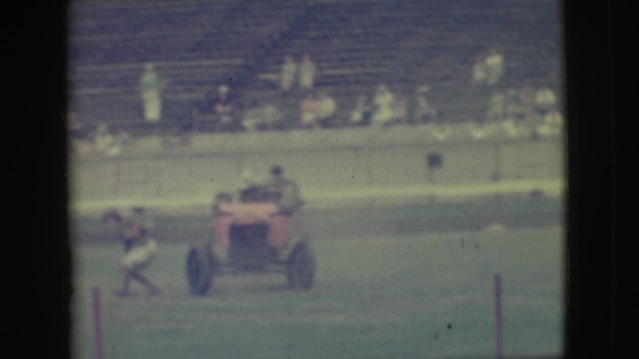 Small rider on a red motorized vehicle crossing a grassy field with spectators in bleachers behind.