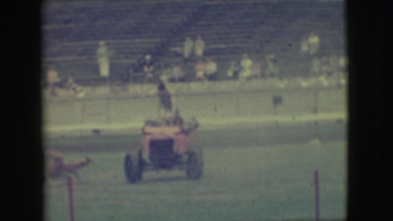 Red motorized vehicle on grass with a rider standing or rising as spectators watch from bleachers.