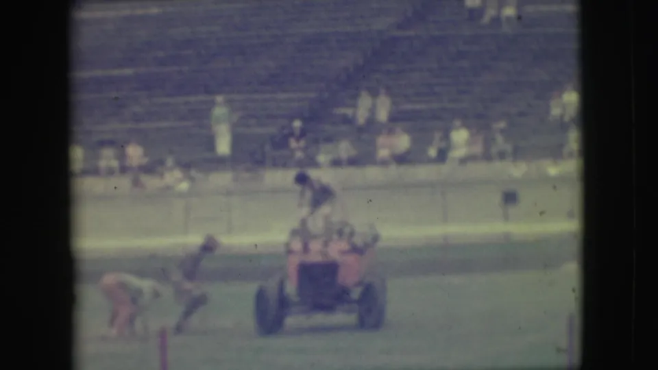 1949 Denver home-movie frame with a person near the small red vehicle on grass and spectator bleachers in the background.