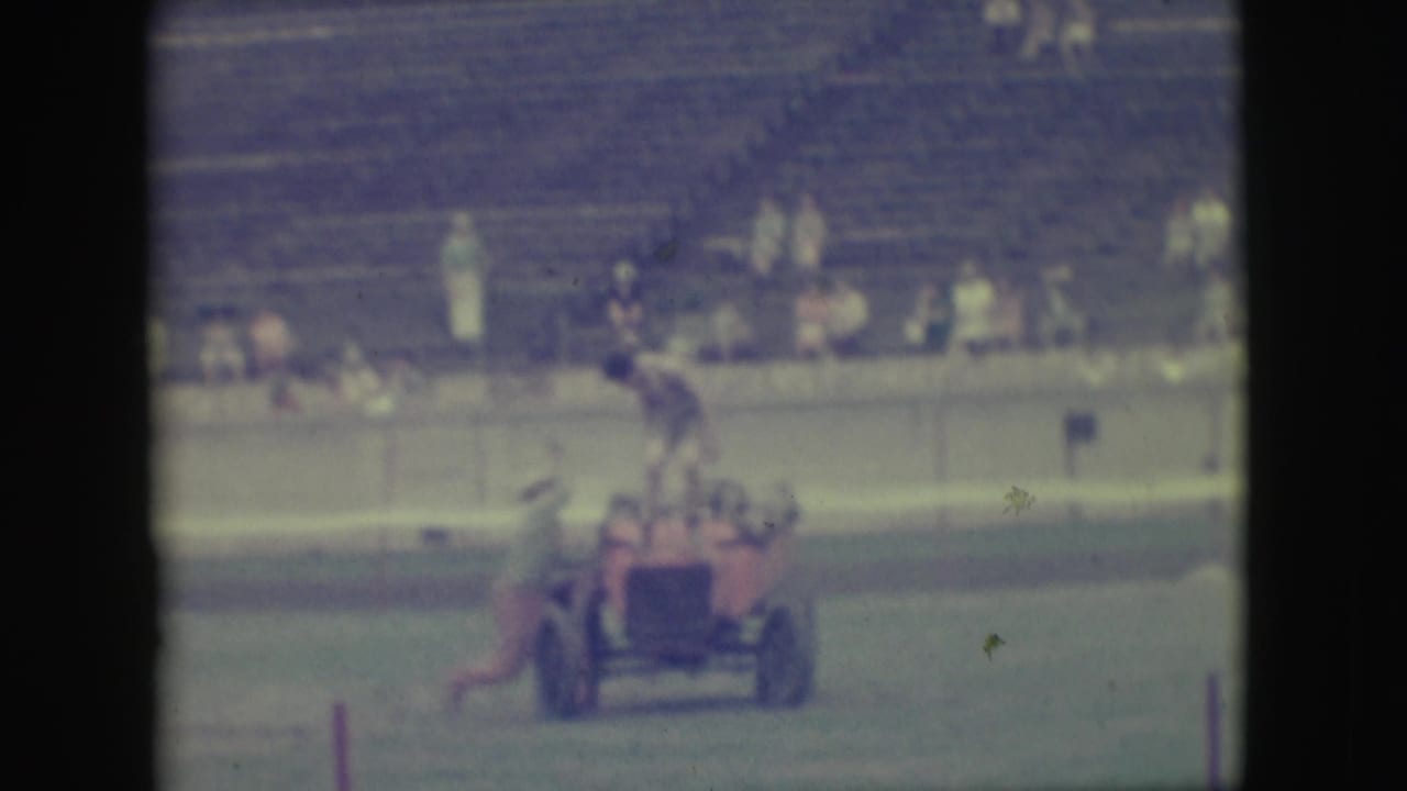 Small red open car with men riding across grass in front of a grandstand.