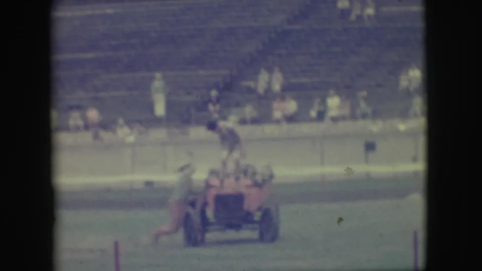 In 1949 Denver home-movie footage, men ride a small red open car on a grassy field before blurred grandstand spectators.