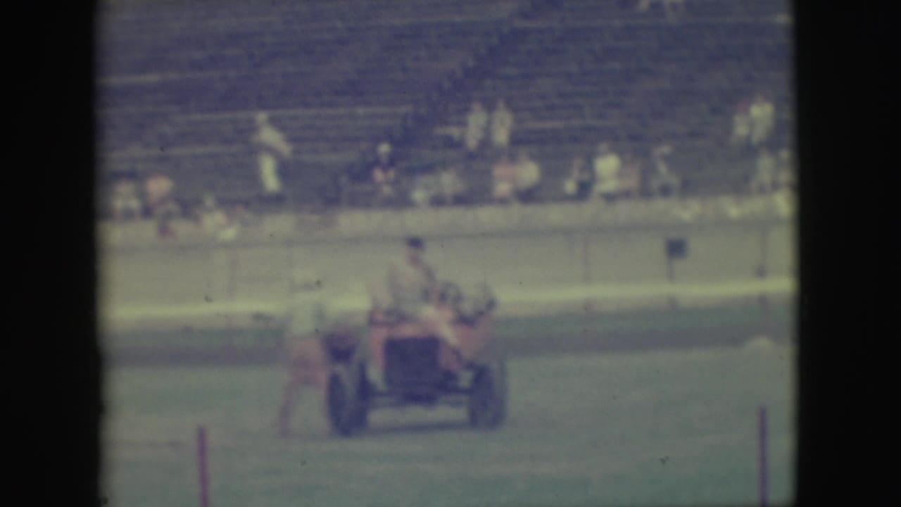 The small red vehicle continues across the grassy Denver field as men ride aboard and spectators sit in the background.