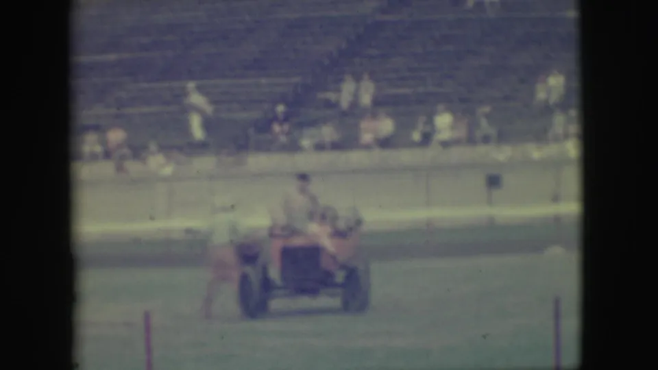 The small red vehicle continues across the grassy Denver field as men ride aboard and spectators sit in the background.