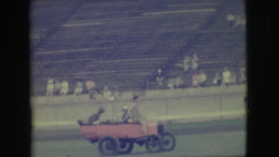 A side view shows the red cart-like car carrying men across the field in front of Denver grandstand spectators.