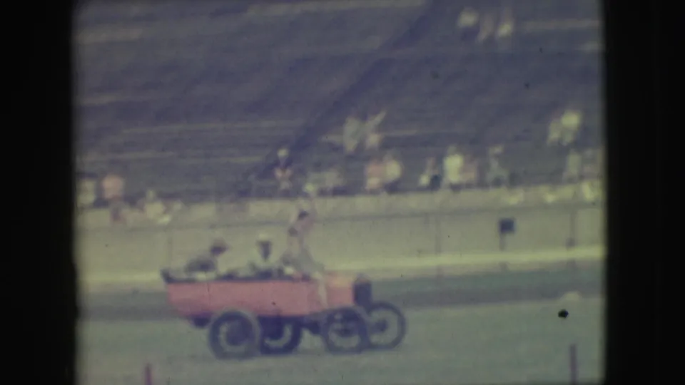 Men ride in the small red open car near the end of the 1949 Denver shot, with distant spectators and tiered seating behind them.