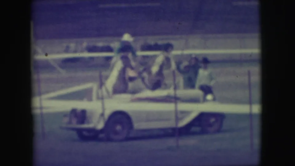In 1949 Denver home-movie footage, a rider balances on two trained horses moving past an arena rail.