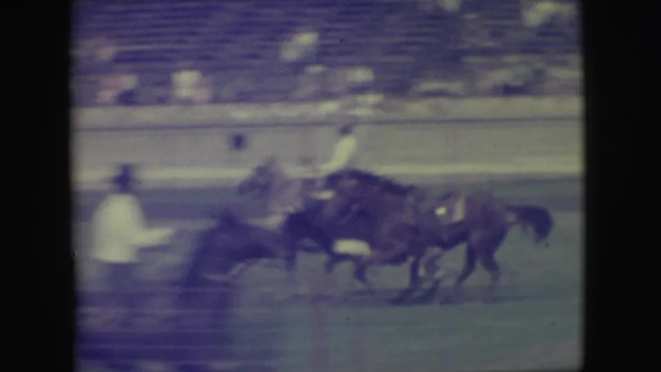 1949 Denver home-movie frame showing a dark horse with rider moving past another person and horse near the arena rail.