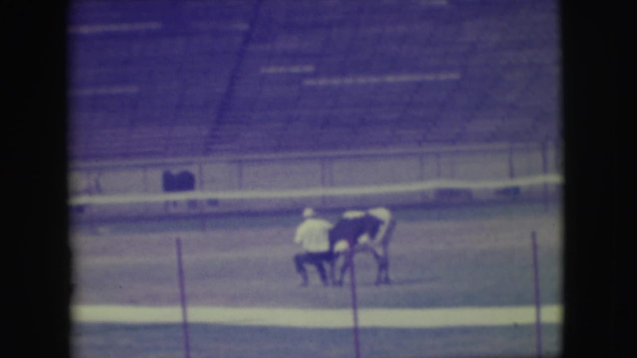In supplied 1949 Denver rodeo footage, adult participants appear near a bull or steer in a dirt arena with empty bleachers behind them.