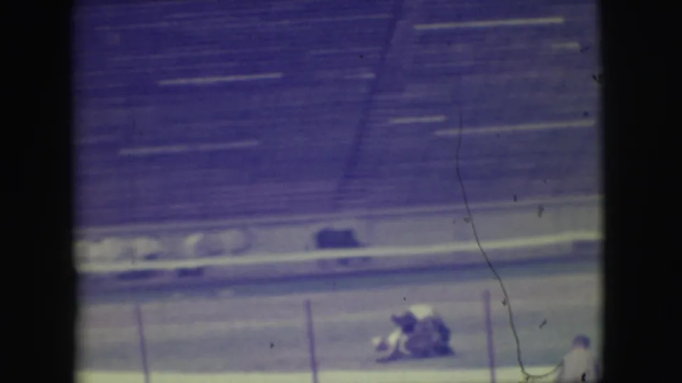 A distant 8mm frame shows the animal and participant low on the dirt in the supplied Denver rodeo sequence, with empty bleachers in back.