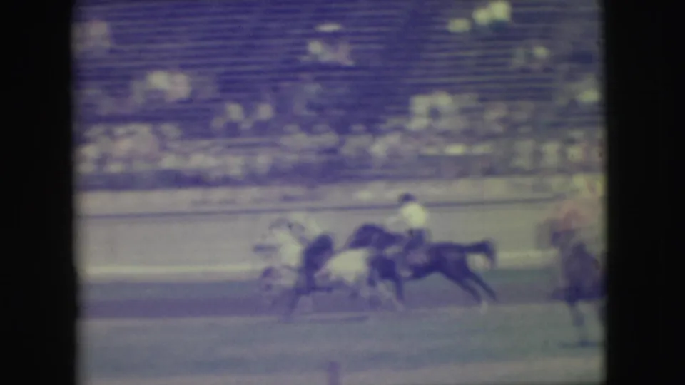 Two mounted riders race along the track in soft 8mm footage, with stadium seating blurred behind them.