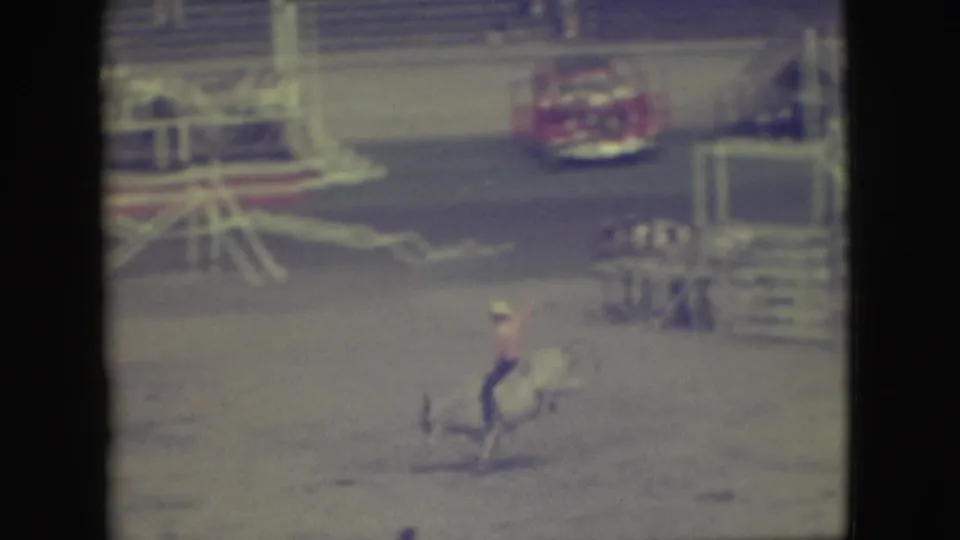 In 1949 Denver rodeo footage, a distant rider in a light hat and pink shirt tries to stay on a bucking bull near arena railings.