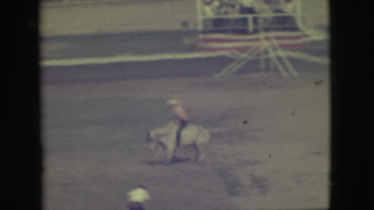 The bull carries the rider across the dusty Denver rodeo arena, with railings and background figures visible in soft 8mm color.