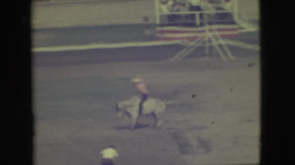 The bull carries the rider across the dusty Denver rodeo arena, with railings and background figures visible in soft 8mm color.