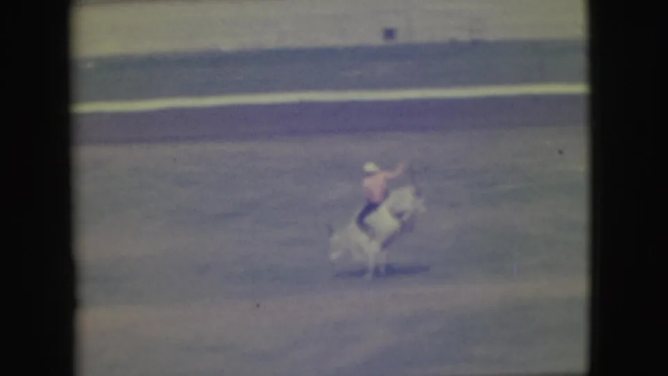 The rider appears thrown upward from the bull in the Denver rodeo arena, captured in a blurred, color-faded 8mm home-movie frame.