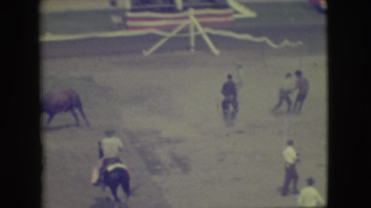 1949 Denver home-movie frame showing a bull and rider in a dusty arena as men watch from nearby positions.