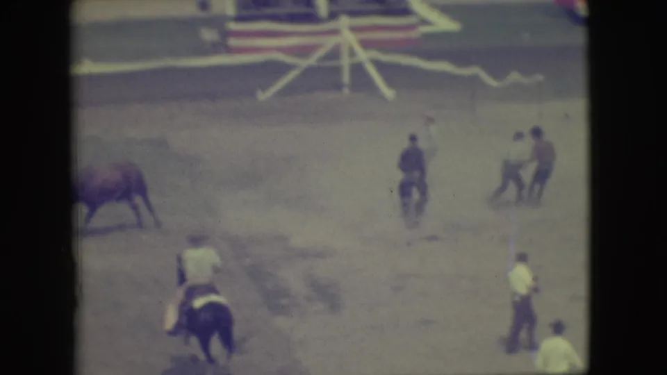 1949 Denver home-movie frame showing a bull and rider in a dusty arena as men watch from nearby positions.