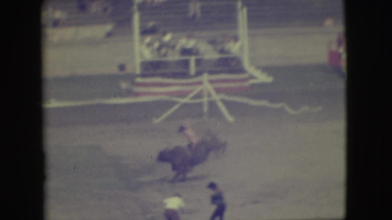 In 1949 Denver rodeo footage, a rider in a red shirt leans off a bucking bull while spectators sit beyond the arena rail.