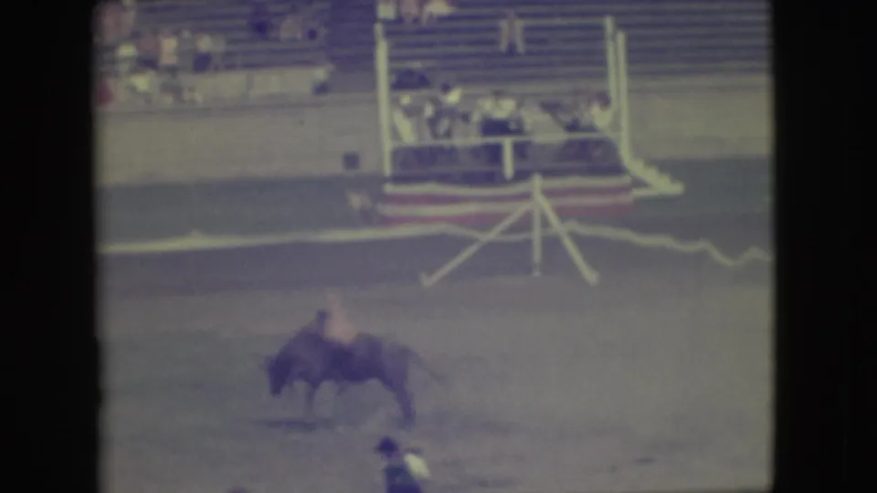 The rider is still mounted on the bucking bull in the Denver rodeo arena, with spectators and railings visible in the background.
