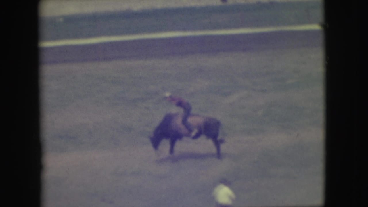 Bull and rider moving across the arena with a person in a light shirt nearby.