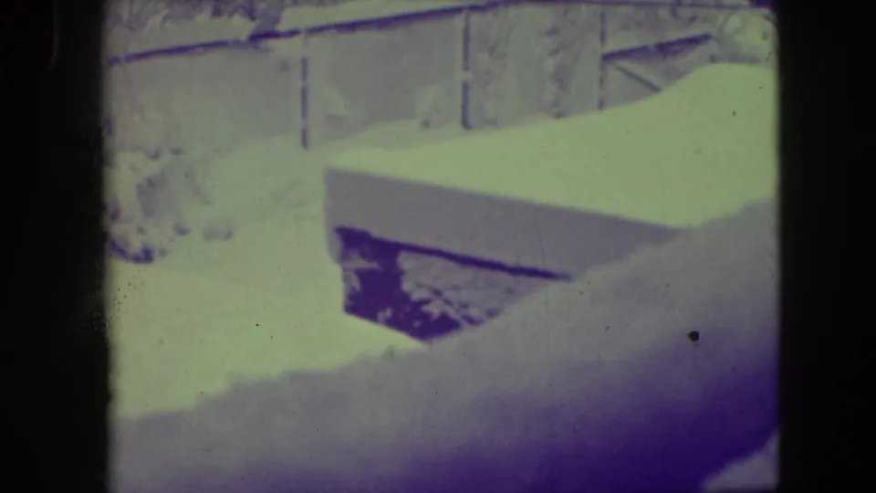 Elmwood Park winter home-movie frame with a brick residential building, porch framing, windows, and snow-covered foreground surfaces in aged 8mm color.