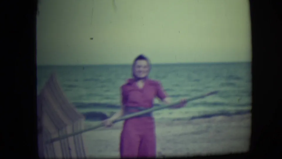 A woman in a pink outfit poses on the Long Beach shoreline, holding a long pole with waves and sand visible behind her.