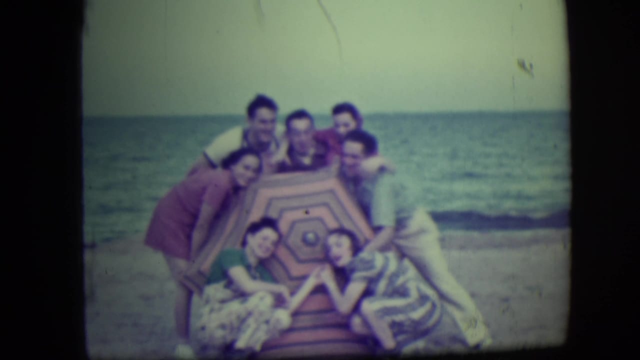Group of young adults posing around a striped beach umbrella near the surf.