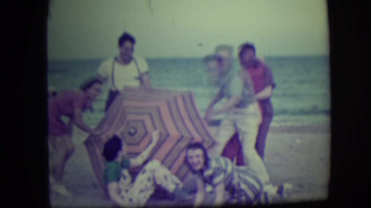 Beach group shifting around a striped umbrella as some people begin to tumble down.