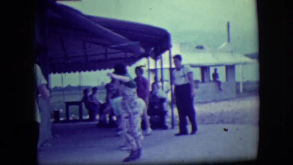 Beachgoers remain near a shaded Long Beach shelter as a person moves through the foreground in soft-focus 8mm footage.