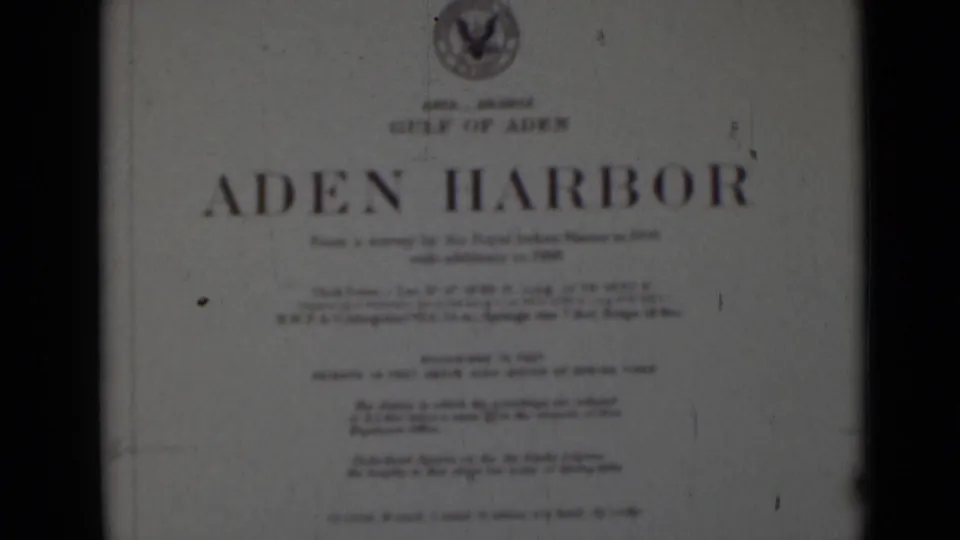 Final frame of the filmed Aden Harbor title card, with blue title lettering, a top emblem, small printed notes, and aged film texture.
