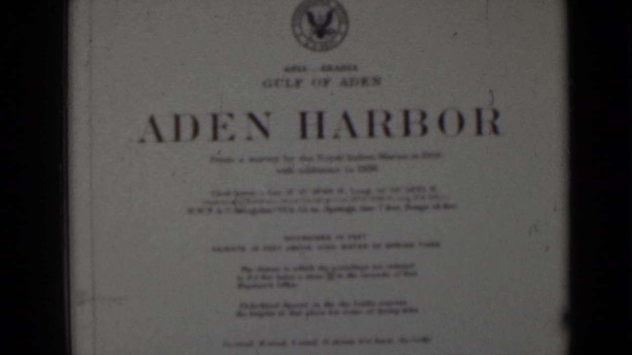 Soft 8mm frame of a printed Aden Harbor title card, with smaller survey text blurred and dark frame borders visible.