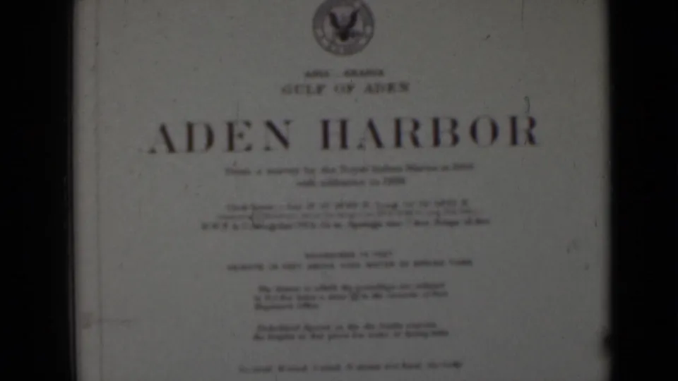 Soft 8mm frame of a printed Aden Harbor title card, with smaller survey text blurred and dark frame borders visible.