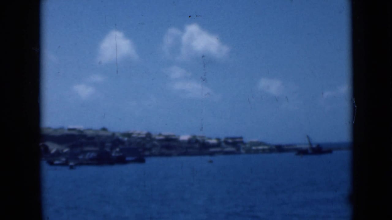 Blue harbor water with a distant shoreline, low hills, small clouds, and a vessel at right.