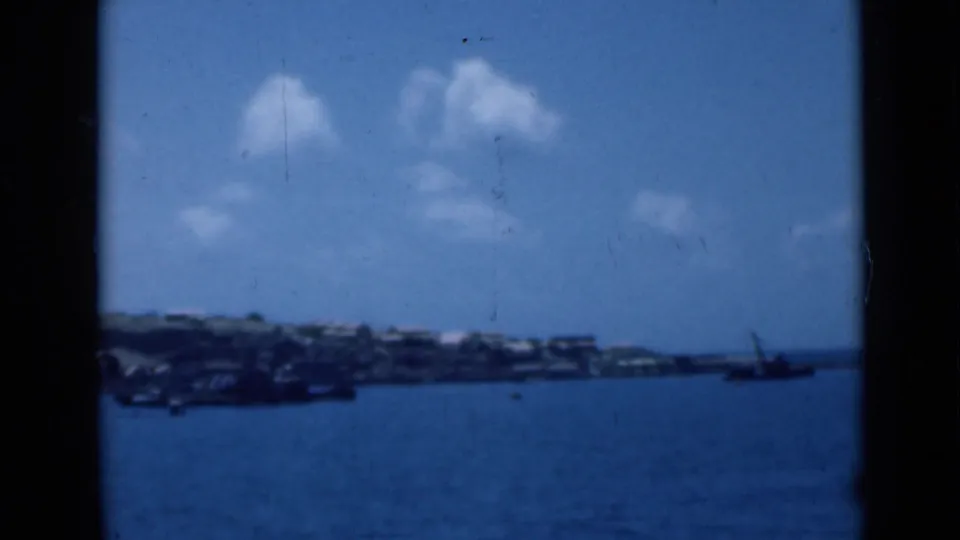 Aden Harbor view from the water, with distant shore buildings, low hills, scattered clouds, and a small vessel near the right edge.