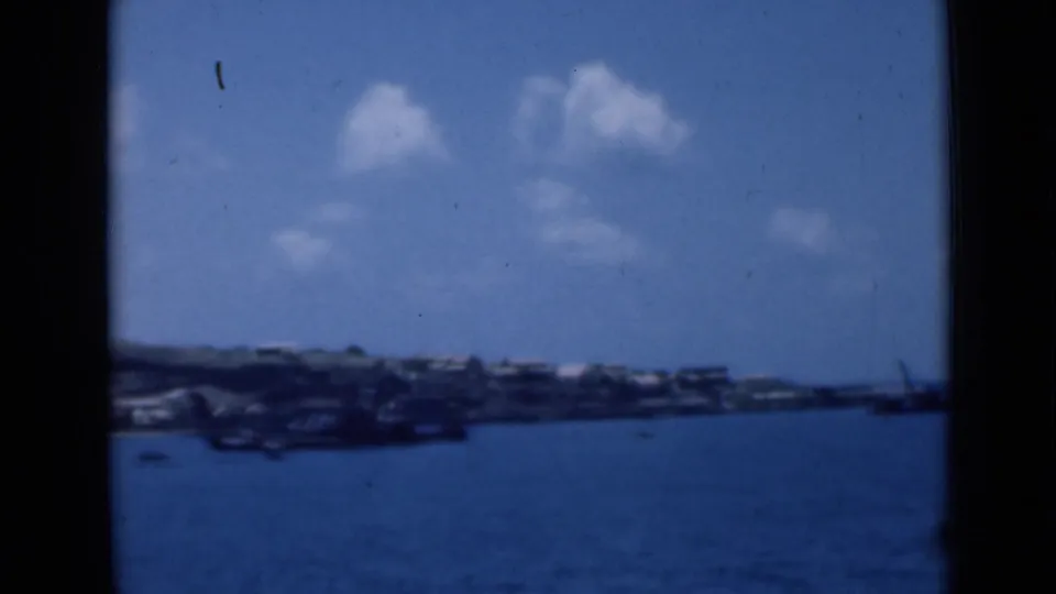 Wide 8mm harbor view showing blue water, distant Aden shoreline, low hills, small boats, and bright clouds under a clear sky.