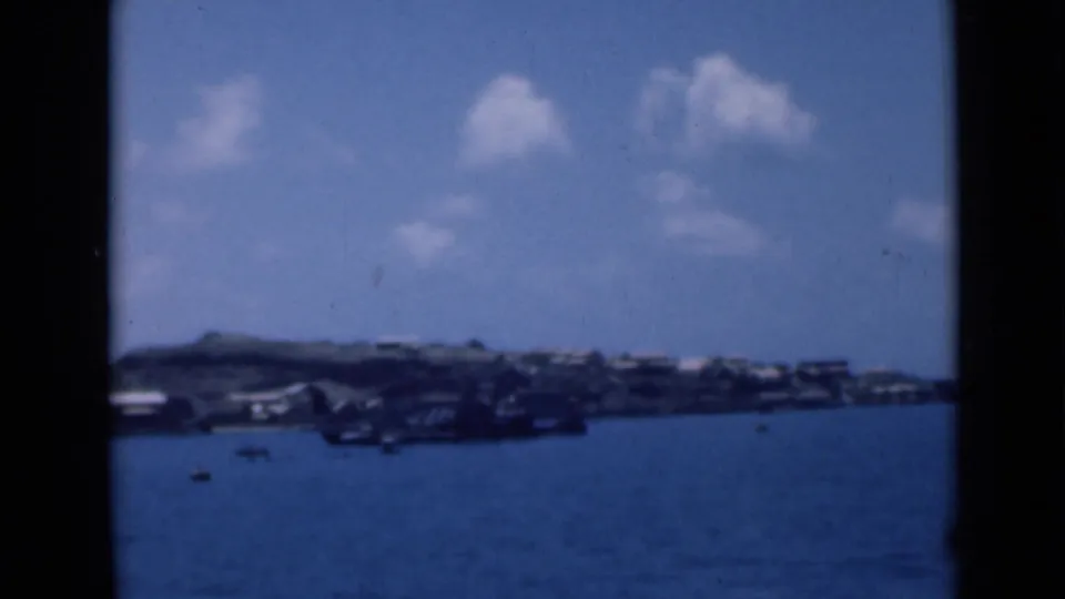 Aden Harbor shoreline seen across blue water, with low hills, small boats, pale waterfront structures, and scattered clouds.