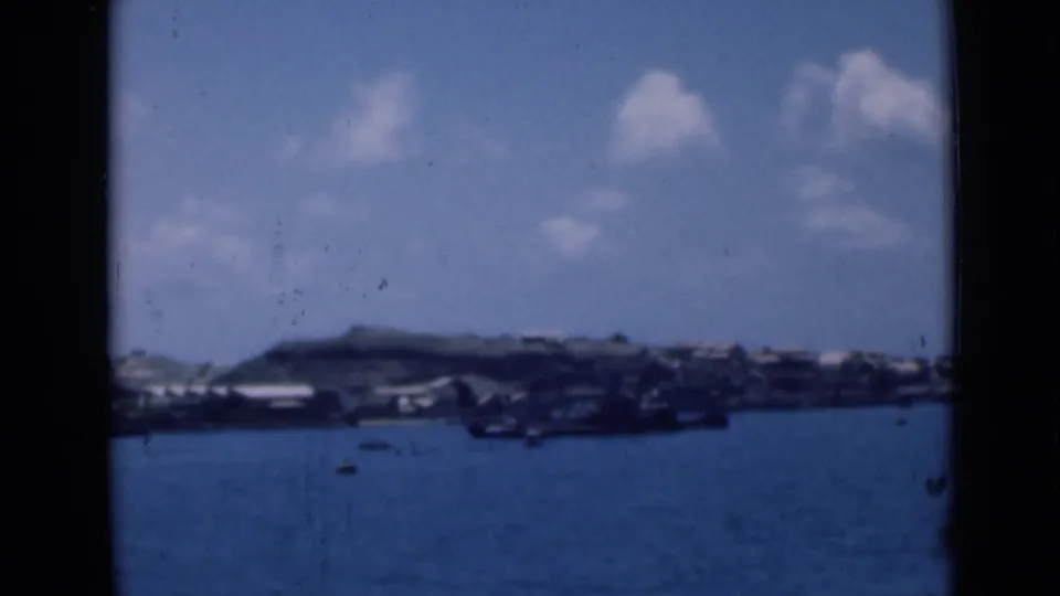 Distant harbor view with clustered shoreline buildings and low hills, captured in soft blue-tinted 8mm home-movie footage.