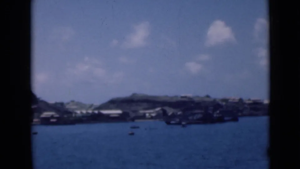 A blue-toned 8mm frame shows small boats on harbor water and low waterfront buildings along a distant hillside.