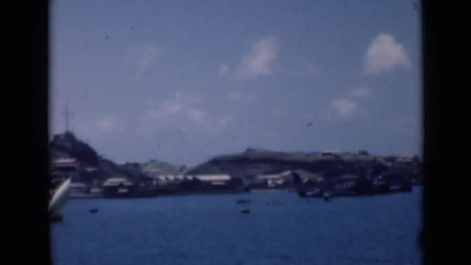 Final soft 8mm view of the distant Aden harbor shoreline, with small boats on blue water and low hills behind buildings.