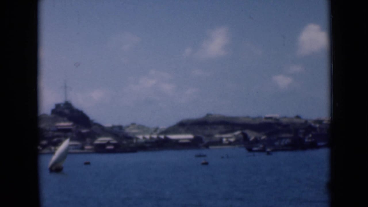 1940s 8mm view of a small sailboat crossing calm water before a distant coastal settlement and low hills.