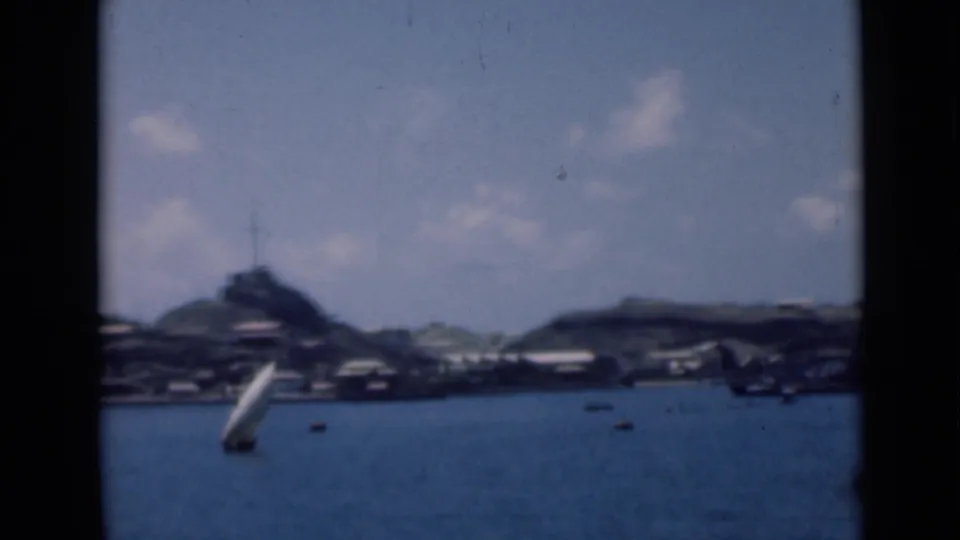 Soft 8mm frame showing a white sailboat on blue water, with low buildings and a hilltop feature in the distance.