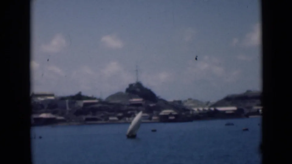 8mm coastal scene with a sailboat in the foreground and distant shoreline buildings below a hilltop structure.