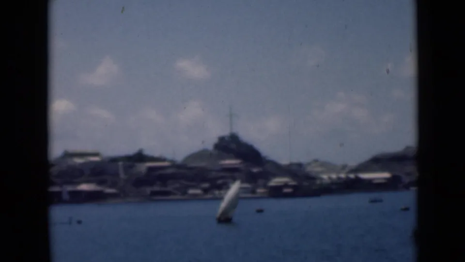 1940s 8mm home-movie view identified with Aden, Yemen, showing a sailboat on calm water before a distant town and hill.