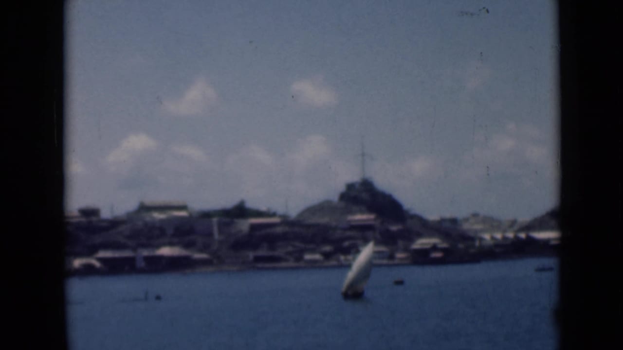 White sailboat on blue water with a distant shoreline, hill, and cloudy sky.