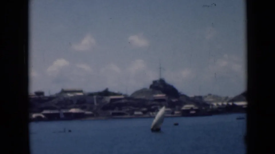 Distant waterfront view from 1946 8mm footage, with a white sailboat moving across the bay below a hilltop marker.